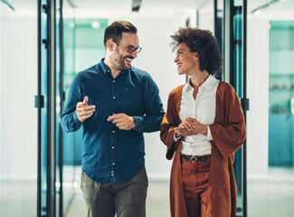 Two coworkers, a white man and a Black woman, walking and talking down a modern office hallway.