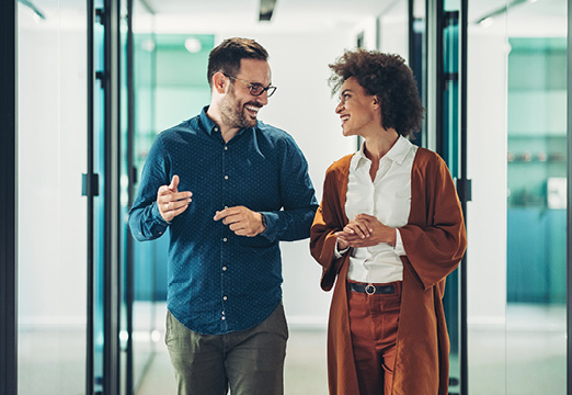 Two coworkers, a white man and a Black woman, walking and talking down a modern office hallway.