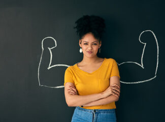 A young woman in a yellow shirt standing against a chalk board with her arms crossed. Muscle arms are drawn on the board behind her.